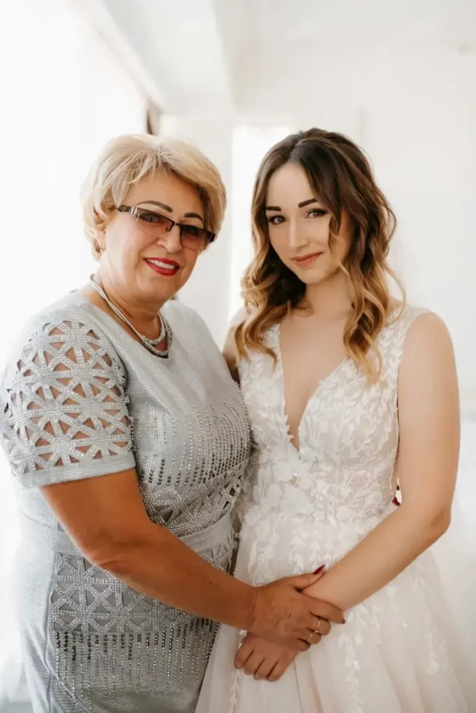 Mother of the bride wearing a stylish, trendy dress standing beside her daughter in a wedding gown, both smiling and posing for wedding photos.